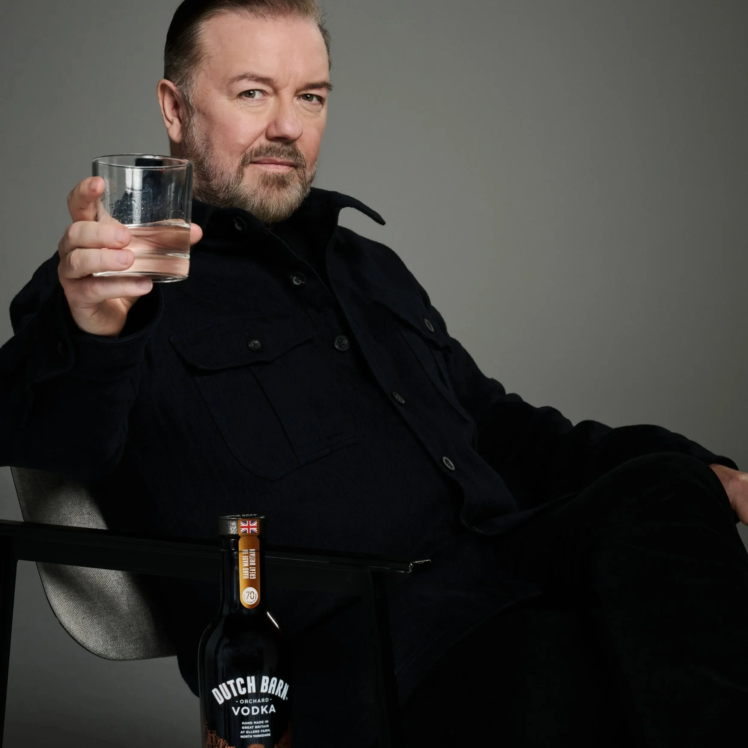 A man with a beard and short hair, dressed in black, seated and holding a glass of clear liquid, with a bottle of Dutch Barn vodka on the table beside him.