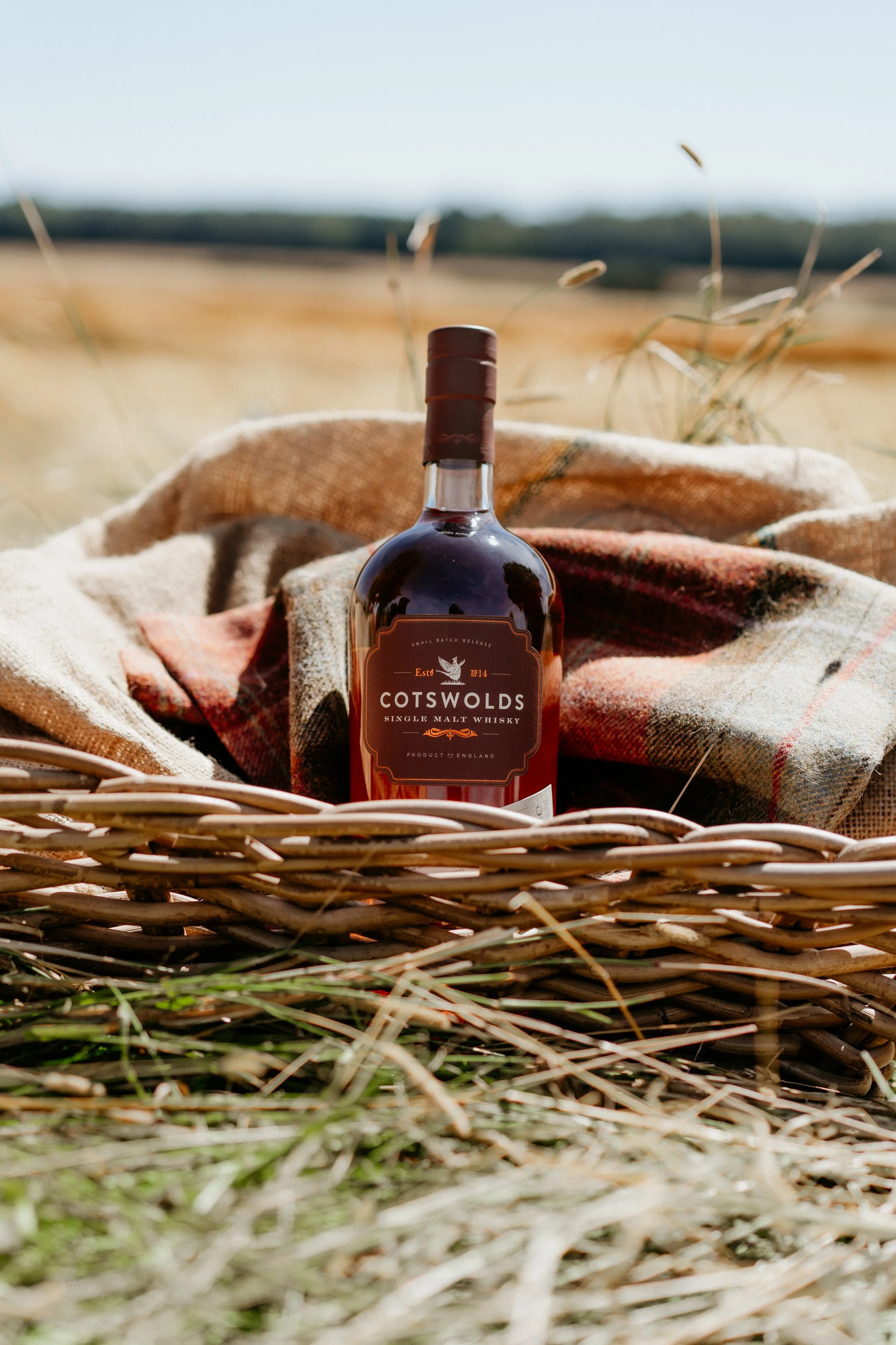 Six bottles of Cotswolds whisky arranged on a wooden platform in a field of wheat with a clear blue sky.
