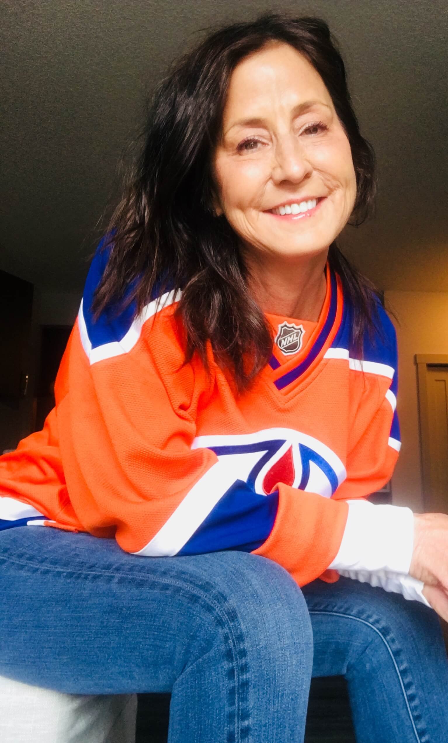 Wholemade Nutrition & Wellness owner, Kathi Decook (NNCP) smiling, wearing an Edmonton Oiler's jersey, sitting on a white chair.