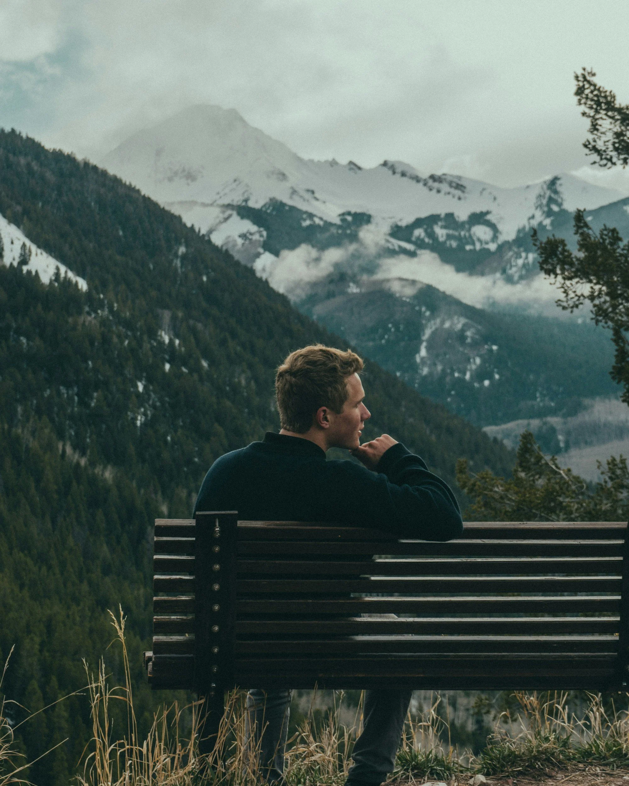 A man sitting on a bench overlooking a mountainous landscape with snow-capped peaks and evergreen trees.