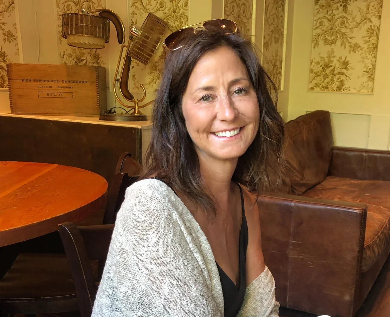 Kathi Decook with shoulder-length brown hair and sunglasses on her head, smiling, seated in a cozy, vintage-style room with floral wallpaper, wooden furniture, and a leather sofa.
