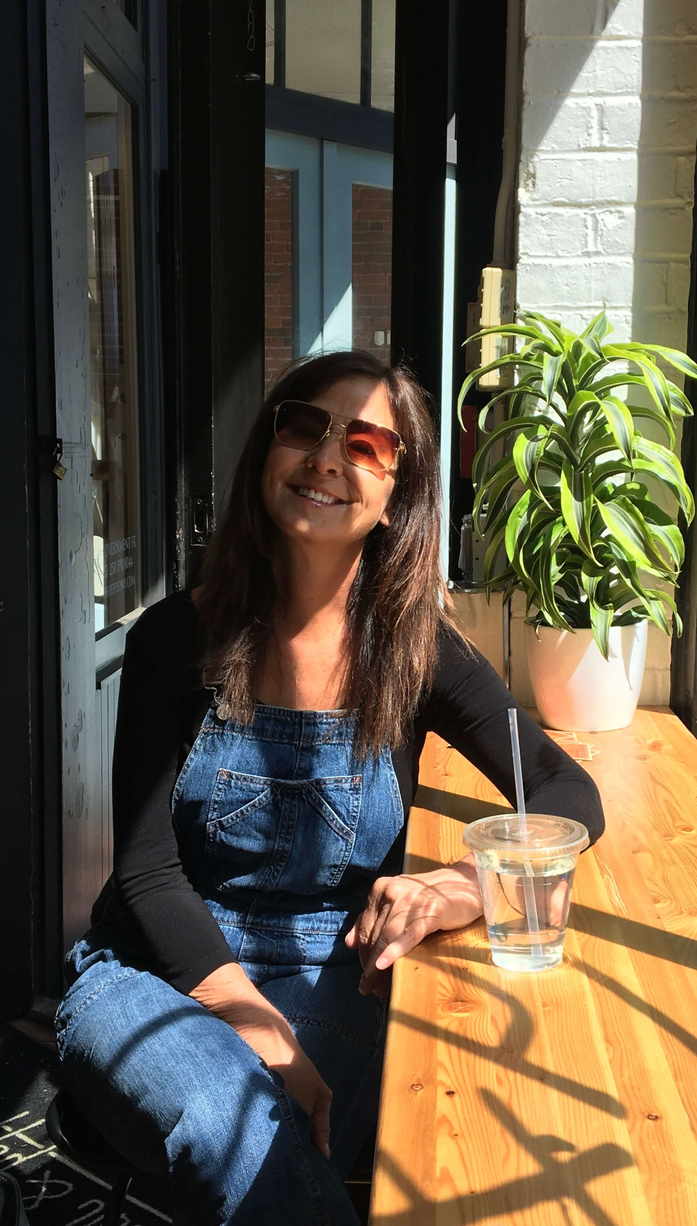Kathi Decook (NNCP) sitting at a wooden table near a large plant, wearing sunglasses and smiling, with sunlight streaming in.