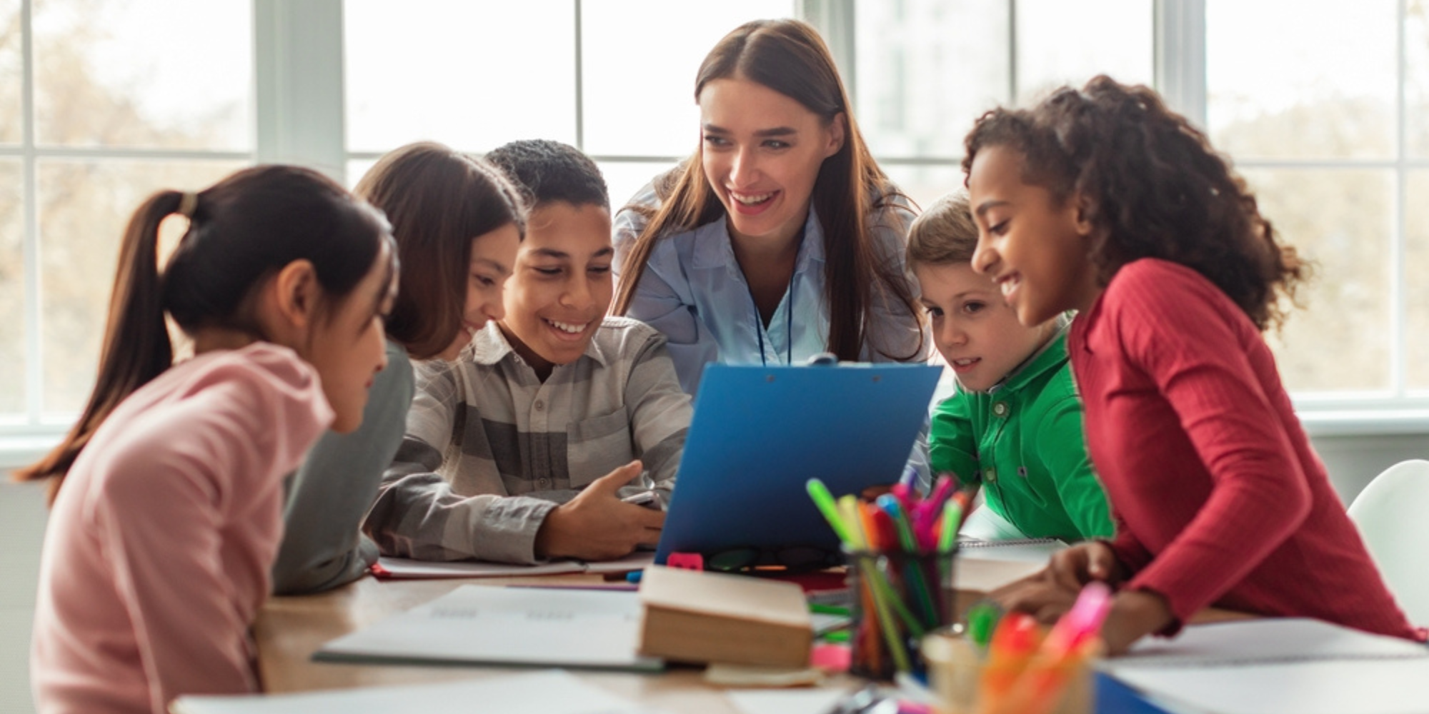 A teacher and five children gathered around a table, looking at a laptop and smiling during a classroom activity.