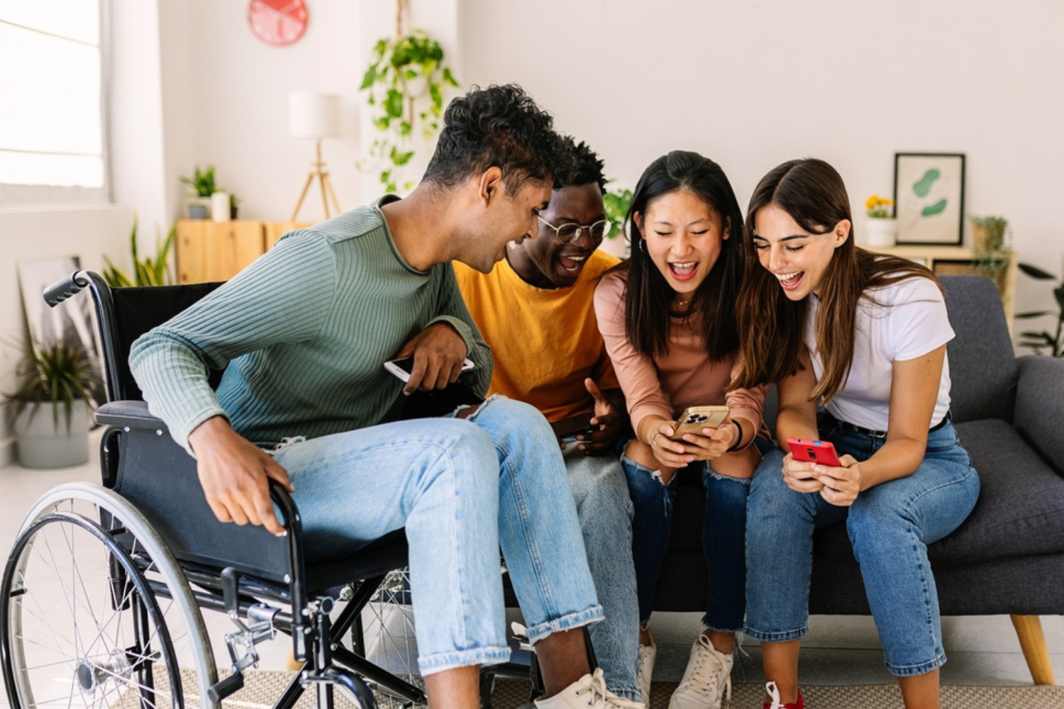 Four smiling young adults sitting closely together on a couch, looking at their smartphones and sharing a moment of joy in a cozy living room.