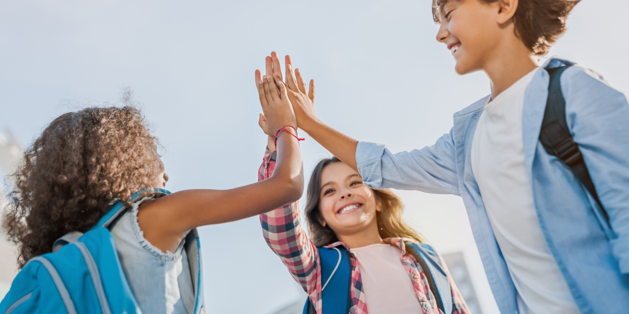 Three smiling children giving each other high fives outdoors on a sunny day.