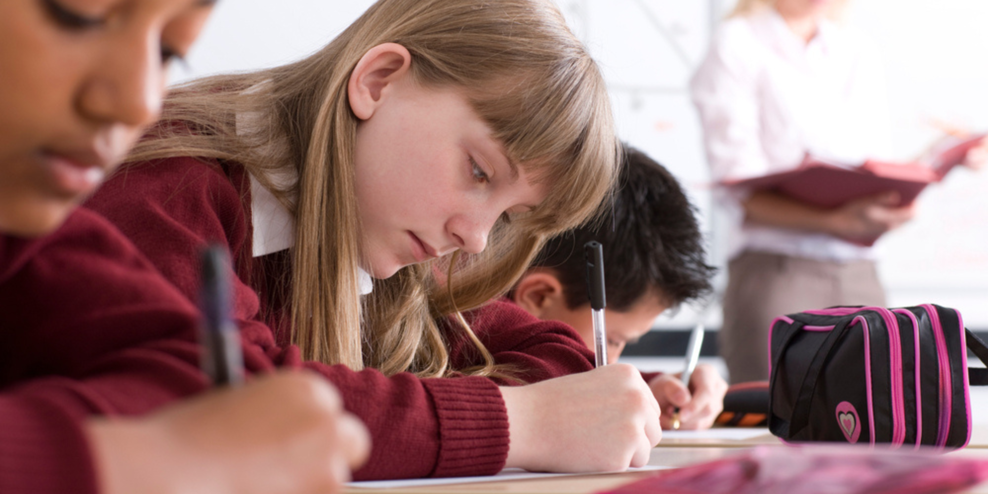Students in a classroom taking a test, with a focus on a girl with long blonde hair in a maroon sweater writing at her desk, and a pink and black backpack nearby.