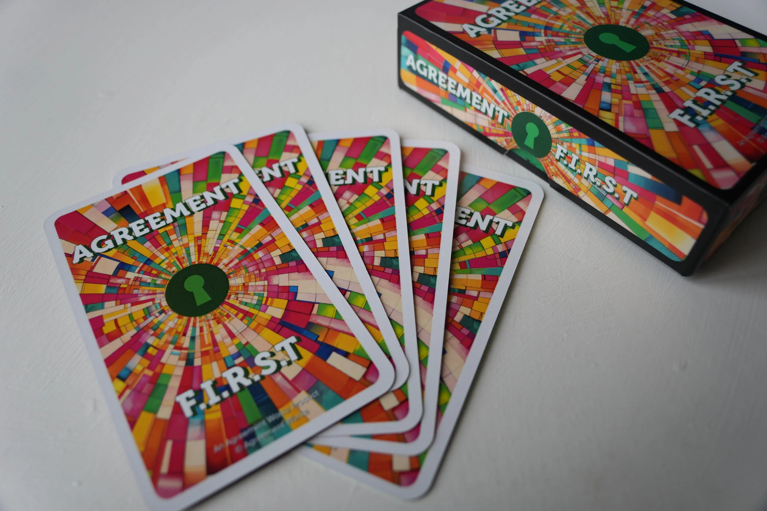 A deck of colorful cards titled 'AGREEMENT FIRST' spread out on a white surface with the box of the game nearby.