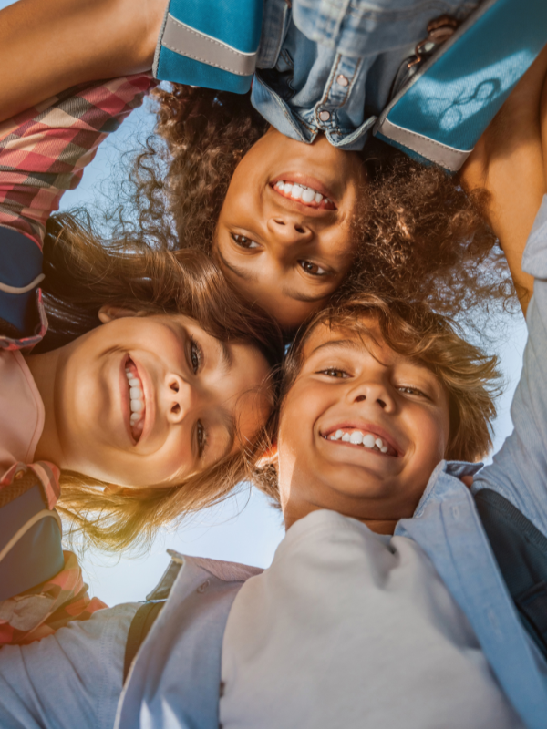 Four children with curly and straight hair smiling and looking down at the camera, forming a circle. The photo is taken from below with a clear blue sky in the background.