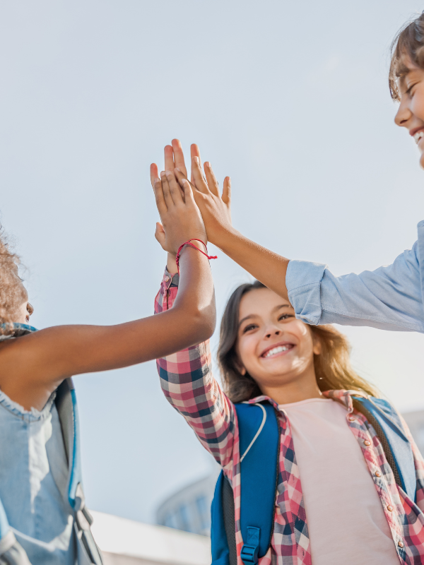 Three children high-fiving outdoors during daytime, smiling and wearing backpacks.