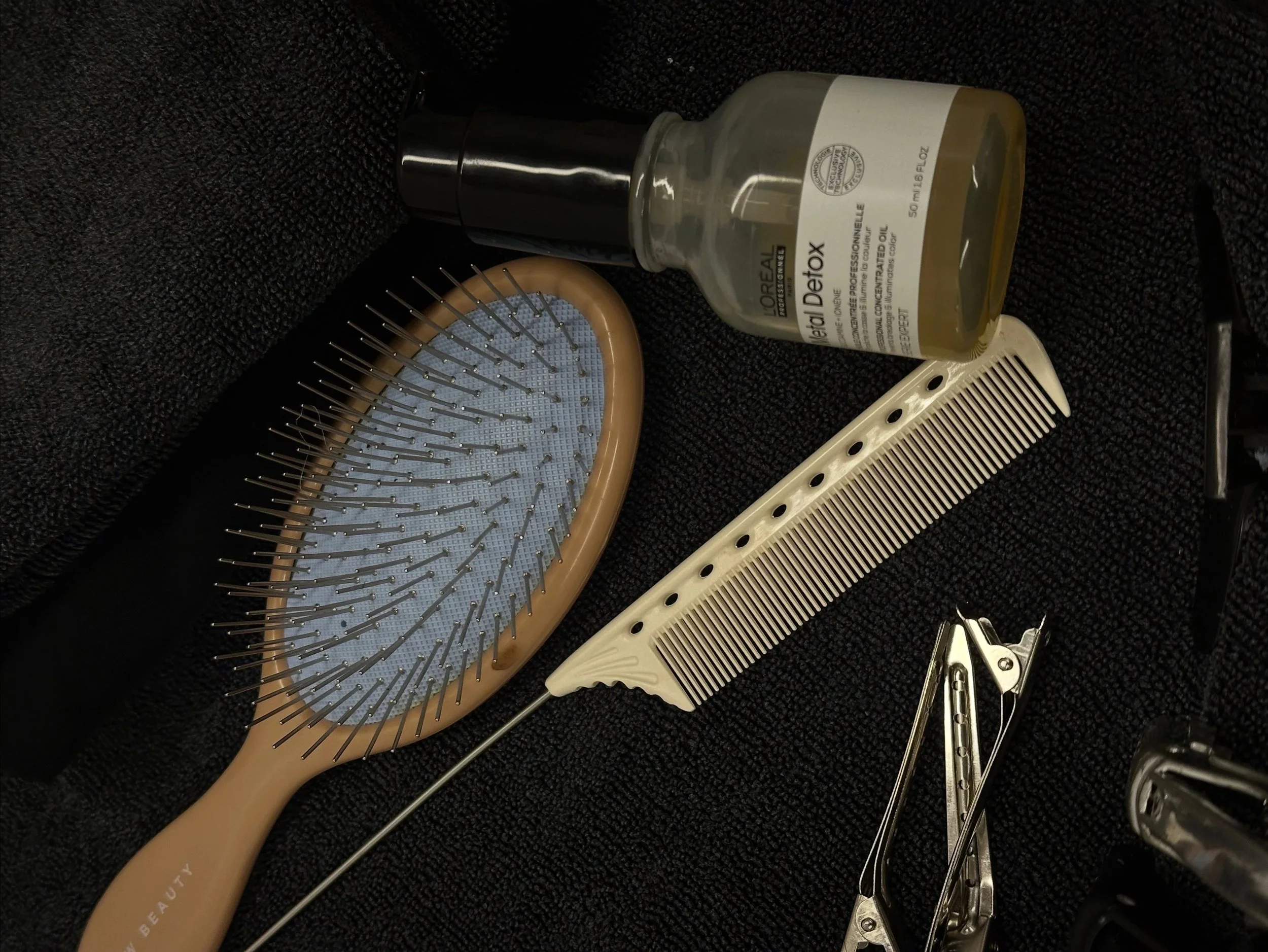 Hairbrush, wooden and white, with metal bristles, and metal hair clips on a black textured surface, along with a bottle of L'Oreal DetoX scalp detox treatment and a small black tool or container.