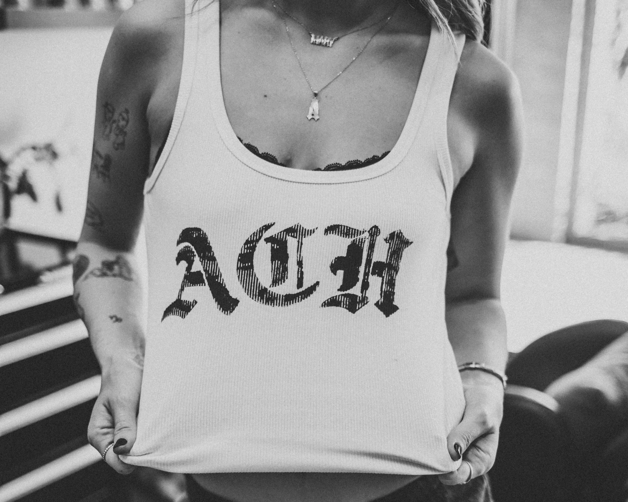 A woman wearing a white tank top with the word 'Hadia' in stylized black lettering holding the bottom of her top with both hands, showing tattoos on her arms and wearing layered necklaces.