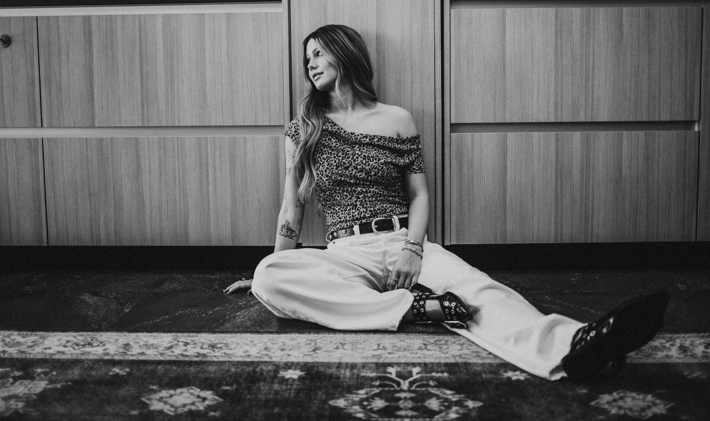 A woman sitting on the floor against a wooden wall, wearing a leopard print top, beige pants, and boots, with tattoos and bracelets, in black and white.