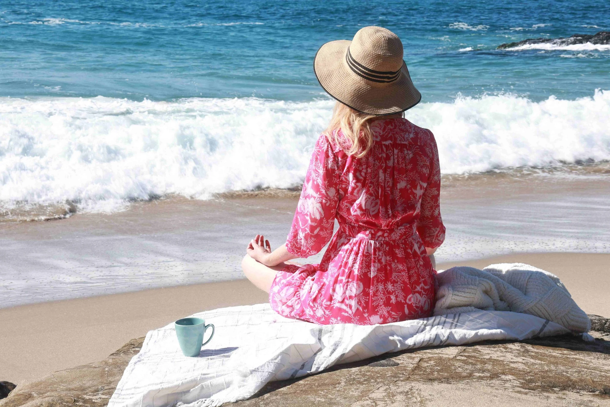 A woman in a pink floral dress and a large straw hat sits cross-legged on a towel on a rock at the beach, meditating near the ocean with waves crashing in the background, and a cup on the towel beside her.