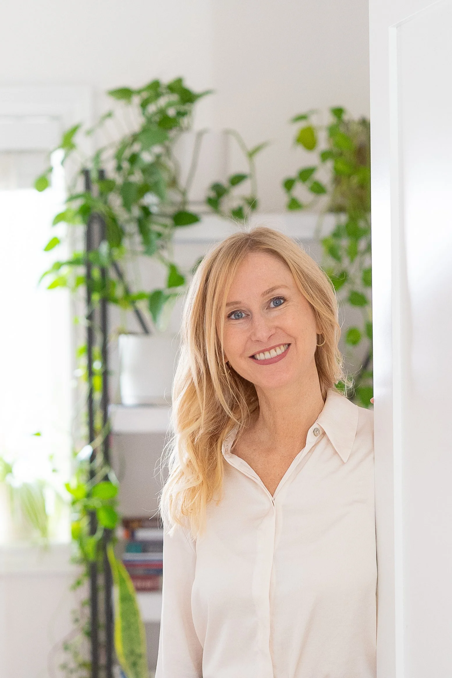 Maria Legault, woman with long blonde hair, wearing a light-colored blouse, smiling and peeking around a doorway in a bright room with green plants in the background.