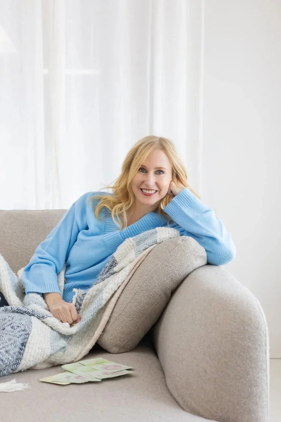 A woman with blonde hair wearing a blue sweater, sitting on a beige couch with a blanket, smiling at the camera, with some banknotes on the couch.