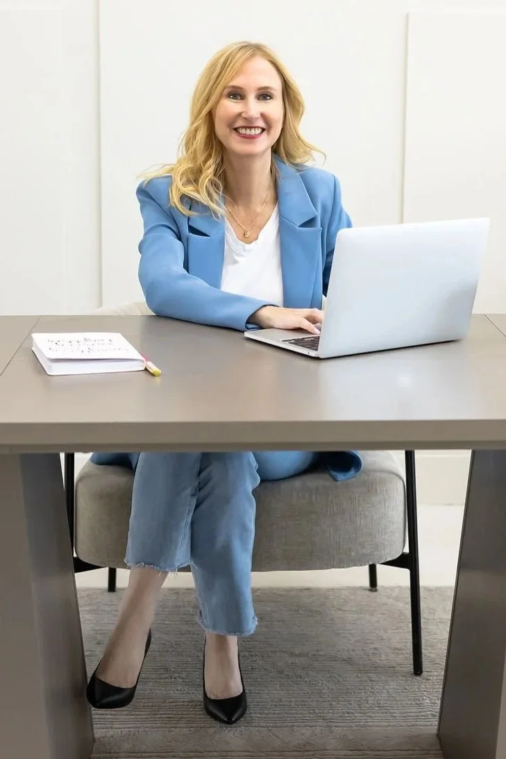 A woman with blonde hair wearing a blue blazer and jeans, sitting at a desk with a laptop, notebook, and pen, smiling at the camera.