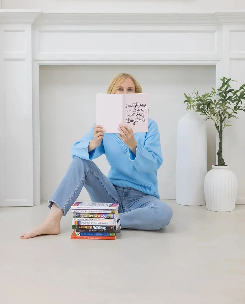 A woman sitting on the floor, holding a sign that reads "everything is coming together," with a stack of books in front of her and a large white vase with a potted plant to her side. She is wearing a blue sweater and jeans, in a bright, minimalist room.
