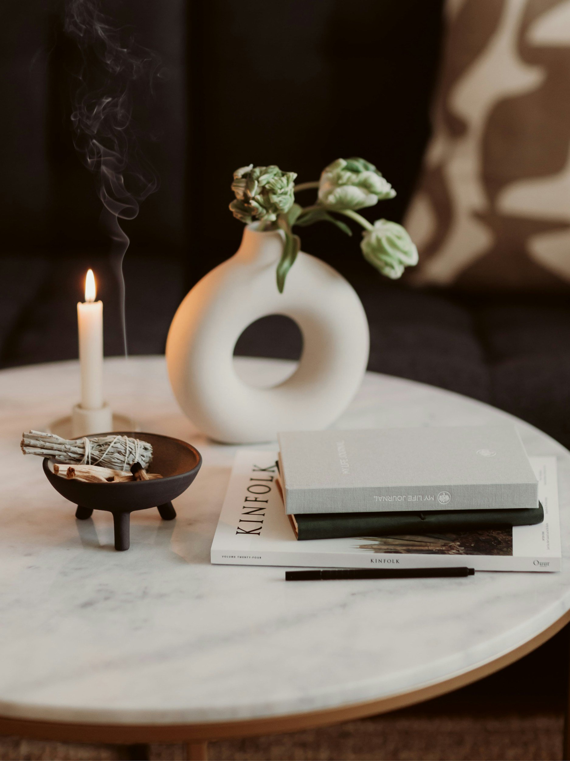 A round marble-topped table with a white ceramic vase containing green flowers, a lit white candle with smoke, a small black bowl with sage bundle and wooden sticks, a stack of books including "Kinfolk," a black pen, and a patterned pillow in the background.