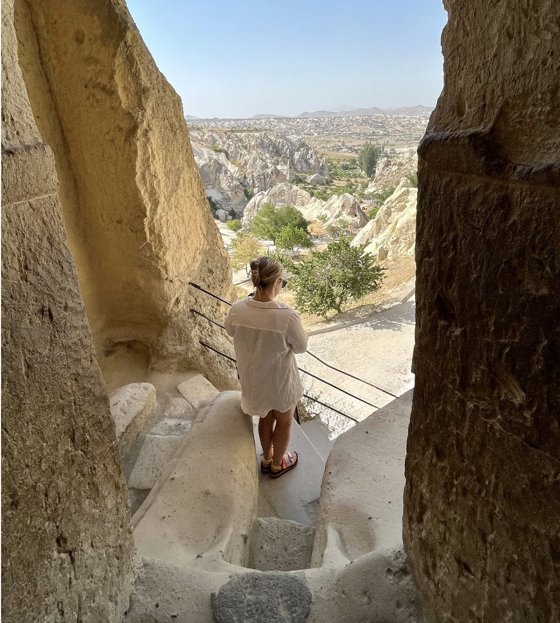 A woman in a white shirt and sandals standing on a rock ledge inside a cave, overlooking a landscape of rocky formations, trees, and distant mountains.