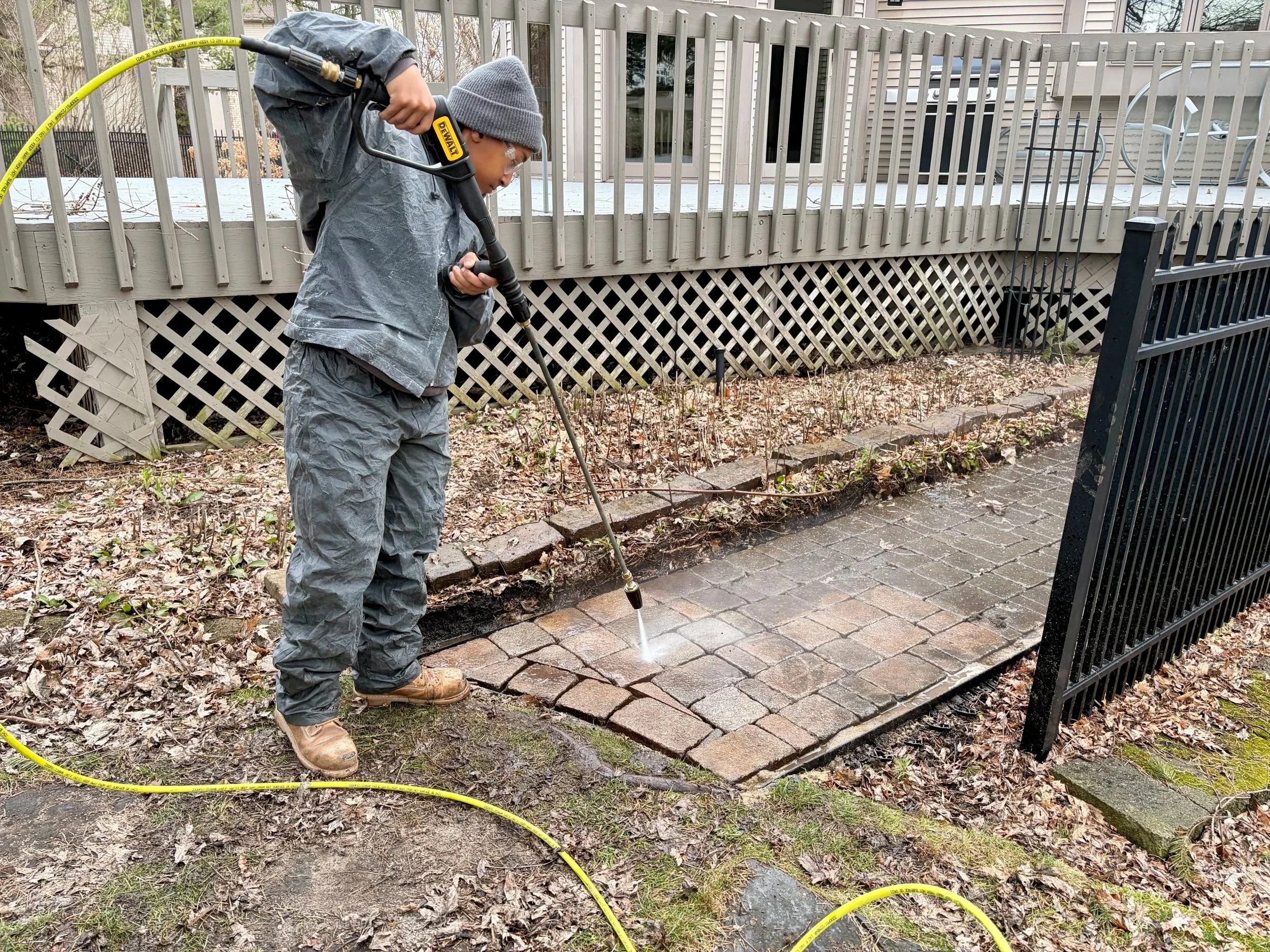 Person wearing gray rain gear and a beanie using a pressure washer to clean a brick pathway outside near a black metal fence.