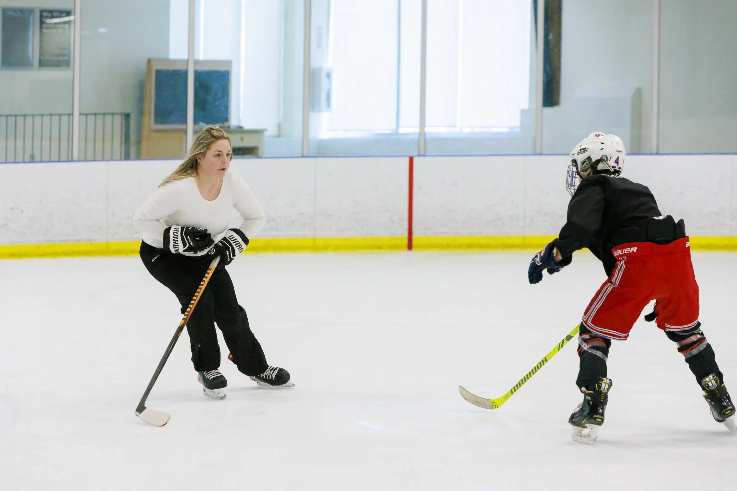 A woman and a child playing ice hockey on an indoor rink. The woman appears to be in a defensive stance while the child is reaching for the puck.