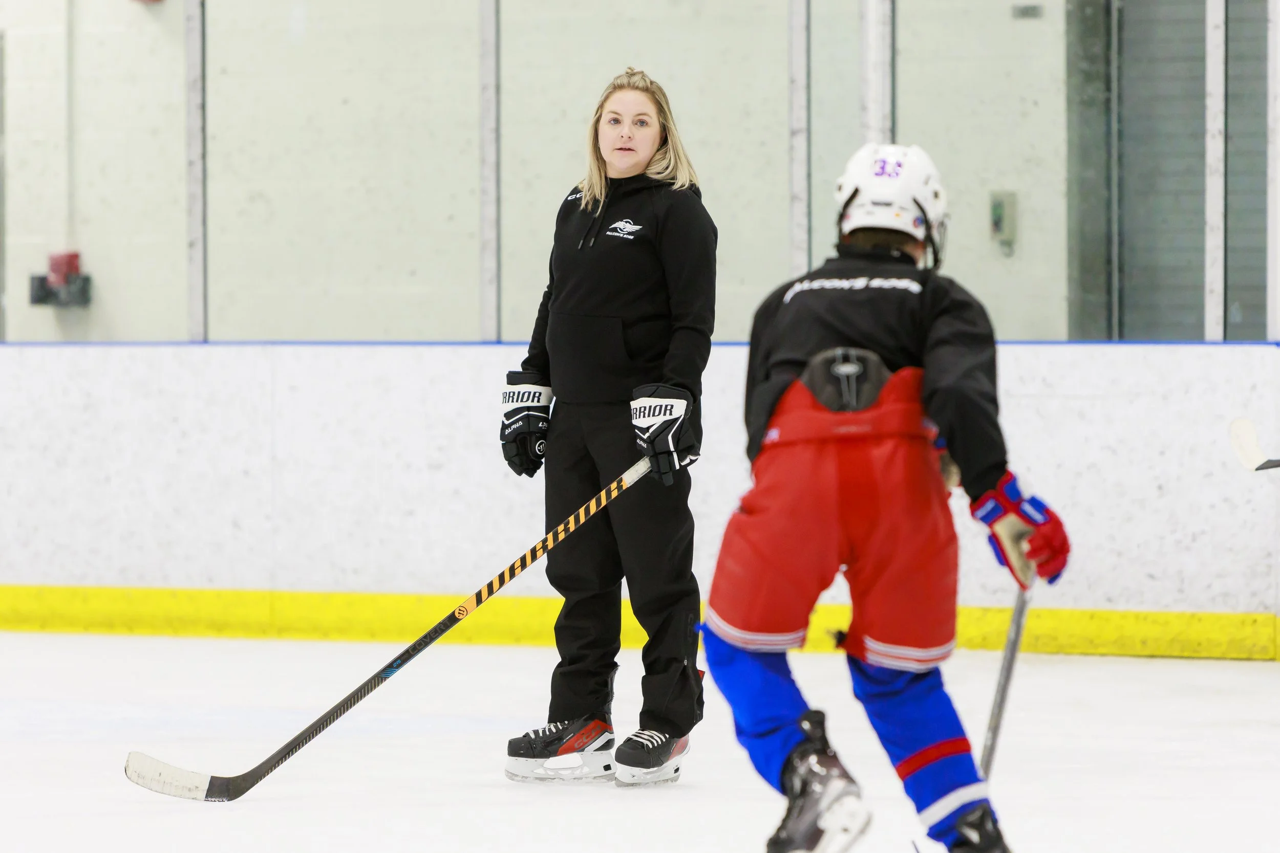 A woman standing on an ice rink holding a hockey stick, wearing black hockey gear, and facing a young child in red and blue hockey attire with a helmet.