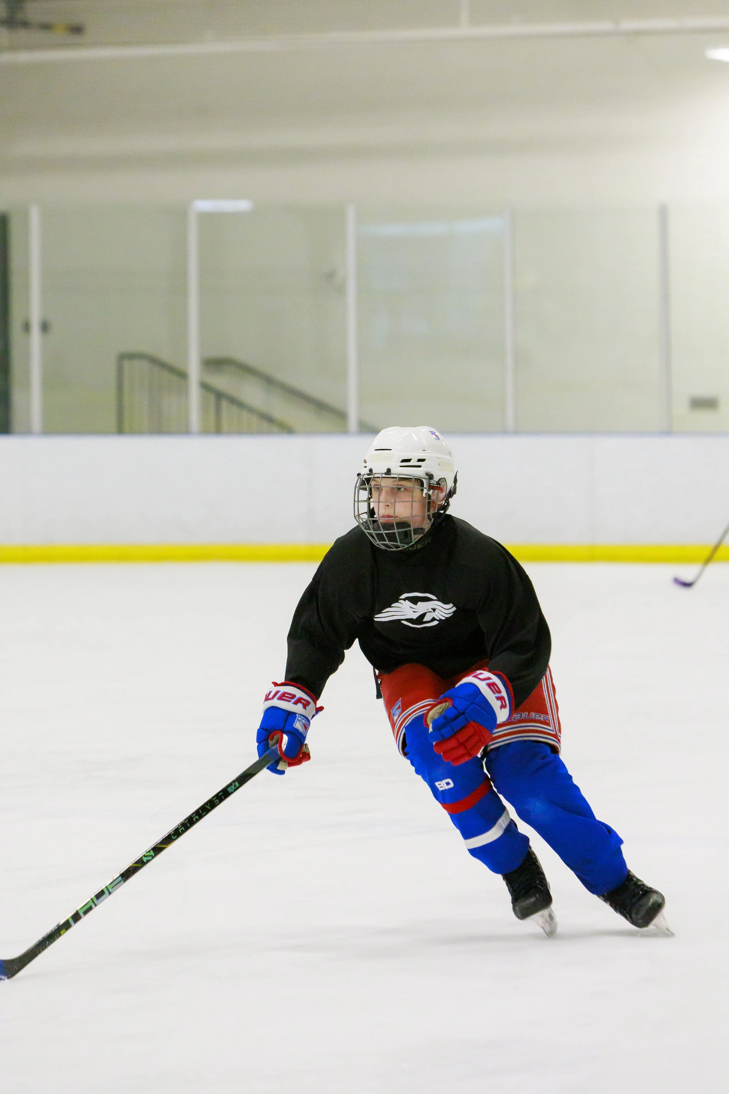 A young ice hockey player in full gear, including a helmet, black jersey, red and blue pants, and gloves, is skating on an indoor ice rink with a hockey stick.
