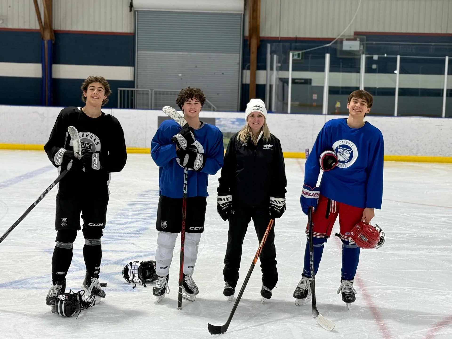 Four teenagers and one woman standing on an ice hockey rink with ice hockey gear and sticks, smiling at the camera.