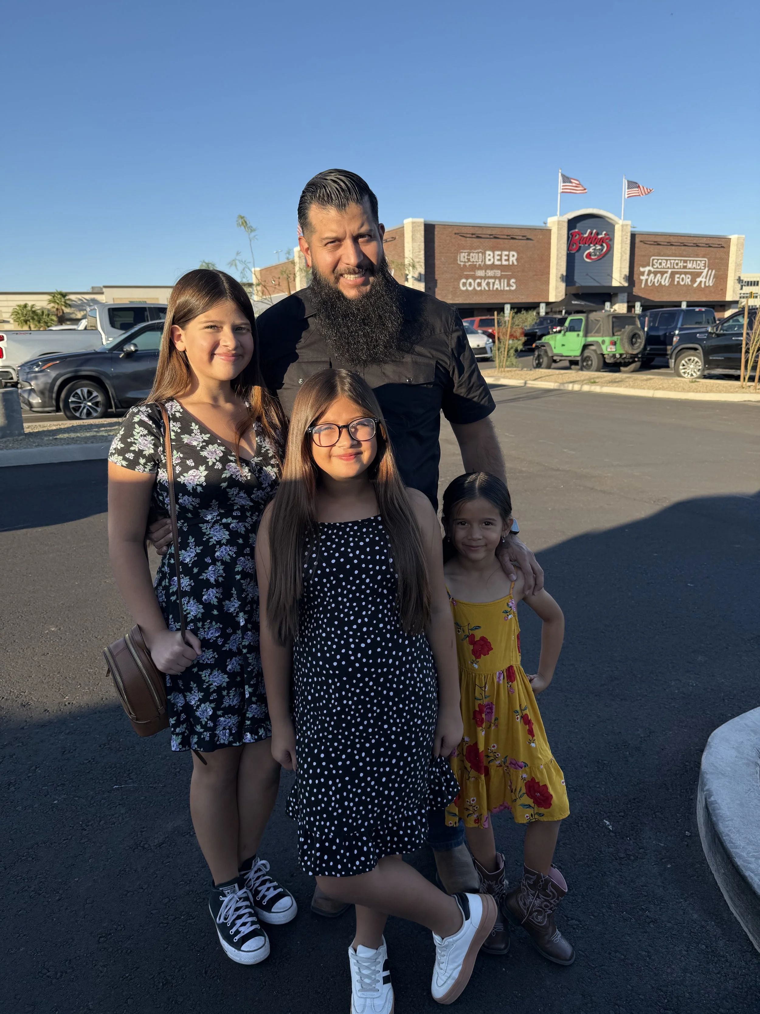 A man with a beard and two women and two young girls standing outside in a parking lot in front of a restaurant called Buddys, with cars parked behind them, under a clear blue sky.
