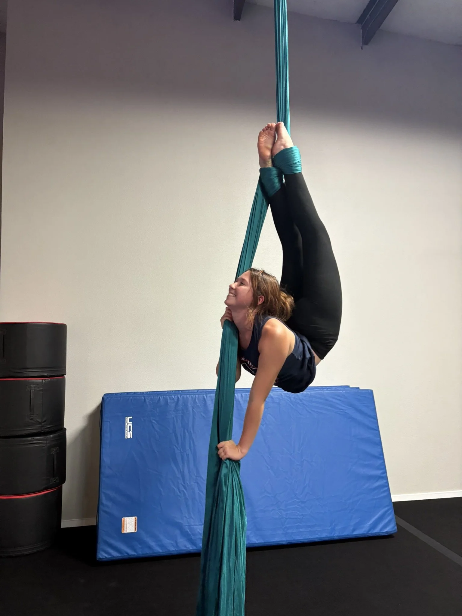 A woman practicing aerial silks, hanging upside down with her legs wrapped around the fabric, while holding with her hands and smiling.