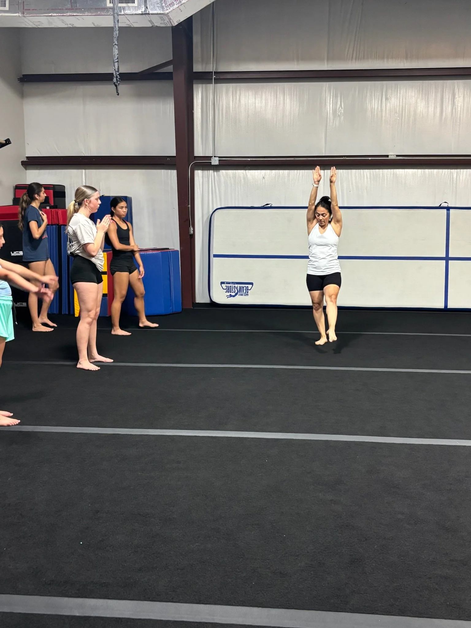 A group of young gymnasts and a coach practicing in a gymnastics training facility. The coach is performing a stretching or warm-up exercise with arms raised overhead, while the gymnasts observe.