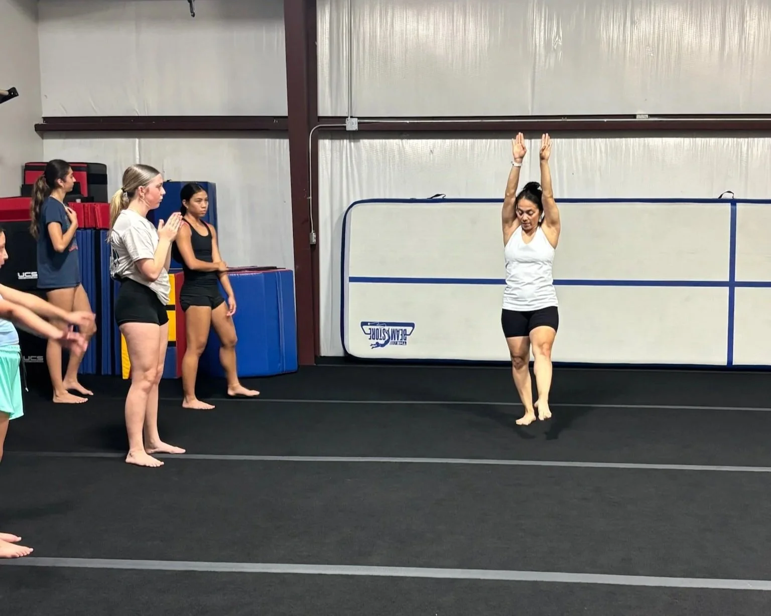 A group of young gymnasts and a coach practicing in a gymnastics training facility. The coach is performing a stretching or warm-up exercise with arms raised overhead, while the gymnasts observe.