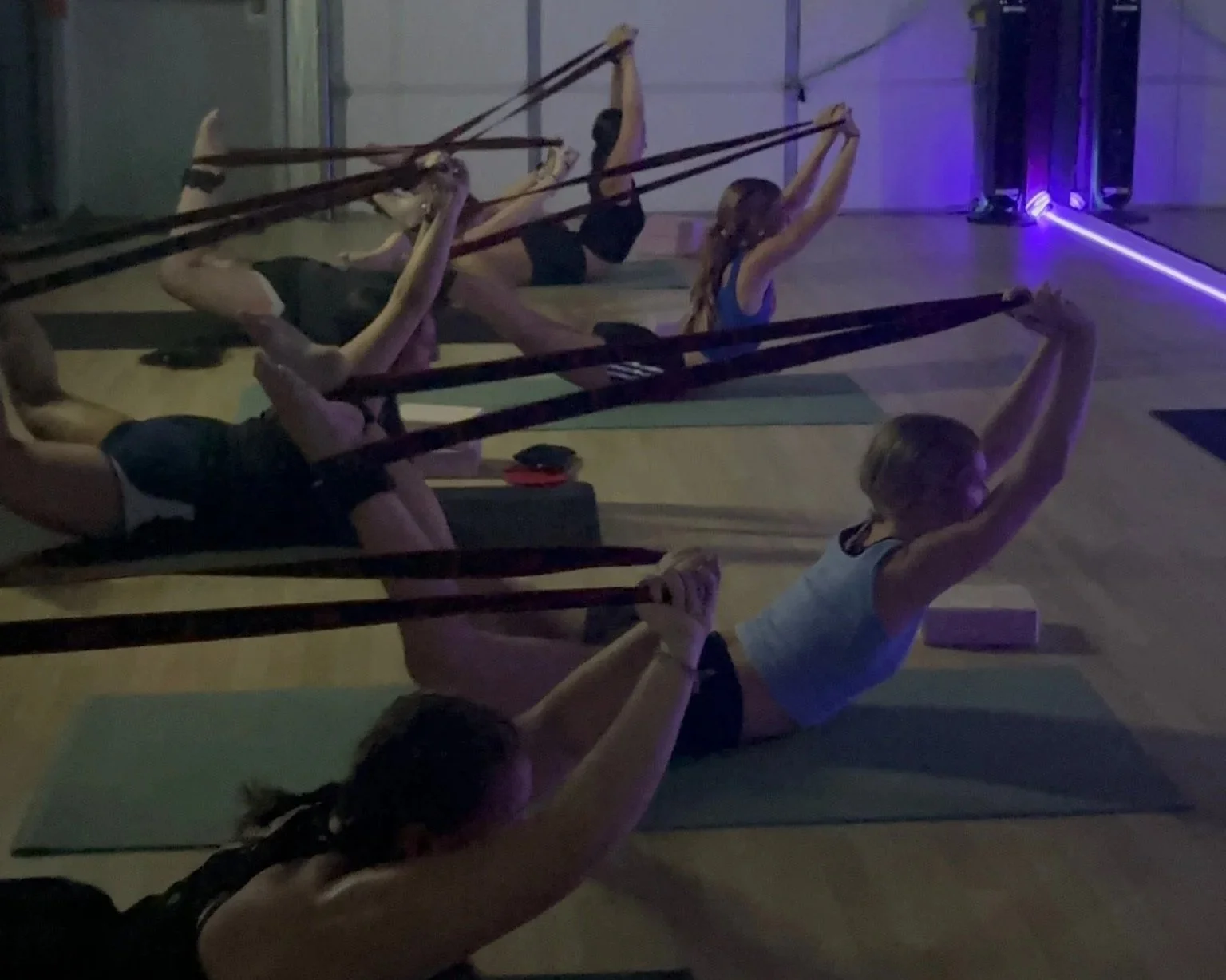 Group of people doing yoga in a dimly lit indoor studio, lying on exercise mats and stretching with resistance bands.