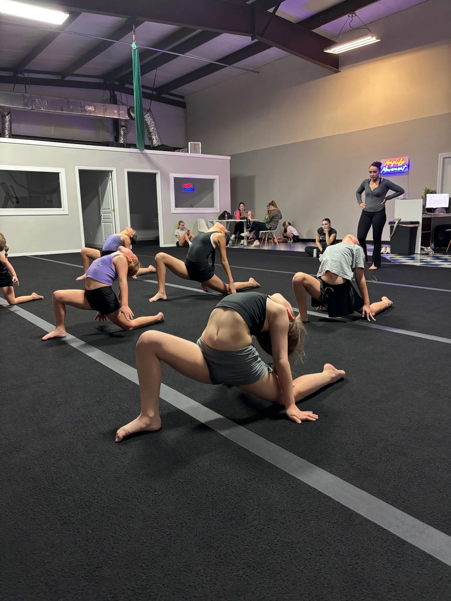 A group of young dancers stretching on the floor during a dance class in a studio.  An instructor stands nearby, observing and guiding the class.
