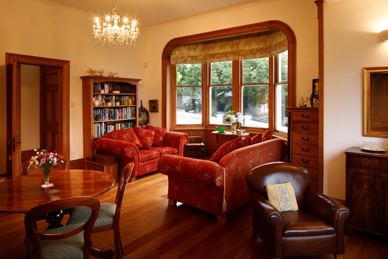 Living room with wooden floors, red sofas, a round wooden dining table with a vase of flowers, a large bay window with trees outside, a chandelier, a bookshelf, and various decorative items.
