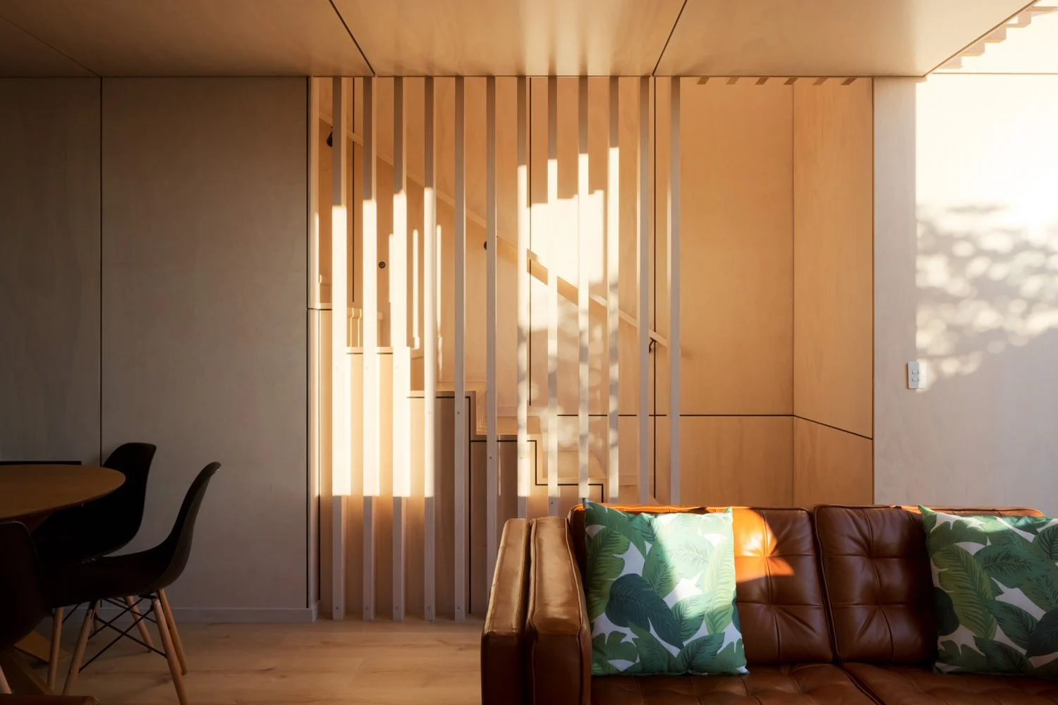 Living room with a brown leather sofa with green leaf-patterned pillows, a wooden dining table with black chairs, a wooden staircase with vertical slats, and warm sunlight casting shadows on the wall and floor.