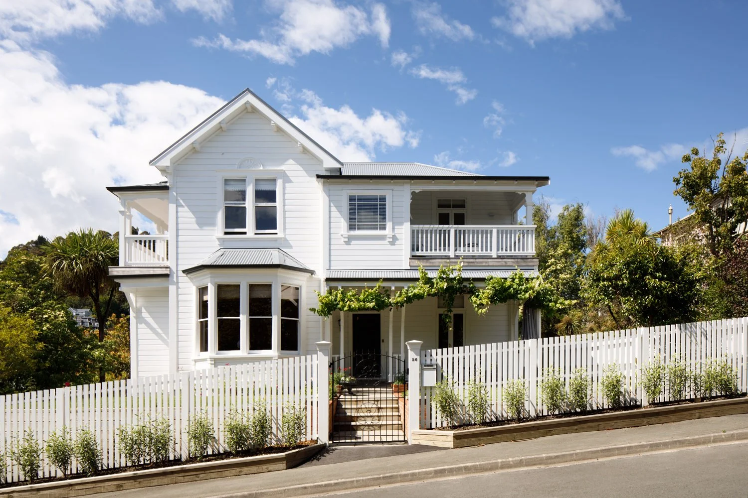 A white, two-story house with a pitched roof, a balcony, a front porch, and a white picket fence. There are trees and shrubbery surrounding the house and a blue sky with some clouds overhead.
