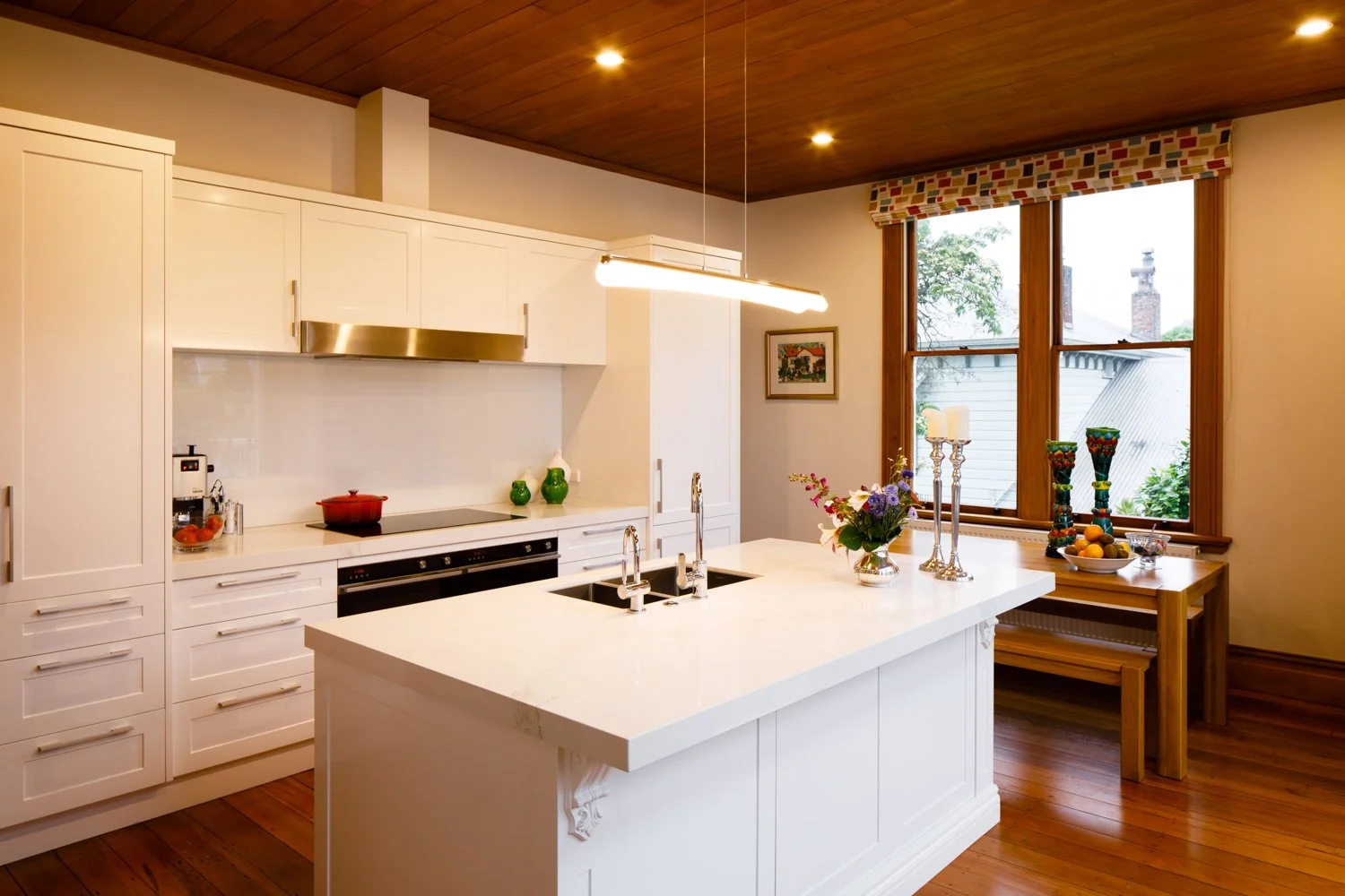 Bright kitchen with white cabinets, island with double sink, wood-paneled ceiling, and large windows with a view of the outside.