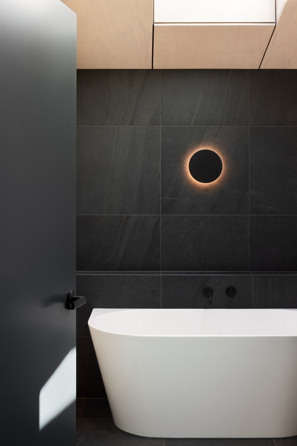 Modern bathroom with black tiled wall, white freestanding bathtub, round mood light on the wall, beige cabinet, and a dark door slightly open with a black handle.