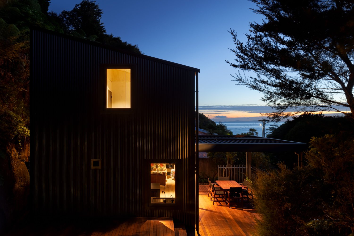 Modern dark house with illuminated interior windows, outdoor wooden dining table with chairs on patio, trees, and ocean view at sunset.