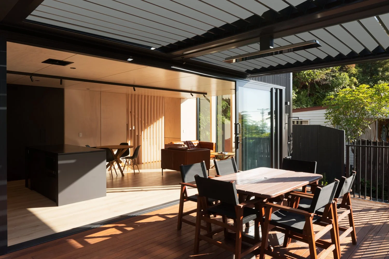 Outdoor deck with a dining table and chairs in front of an open interior living space showing a brown sofa and a wooden wall with vertical slats, with trees and a fence in the background.