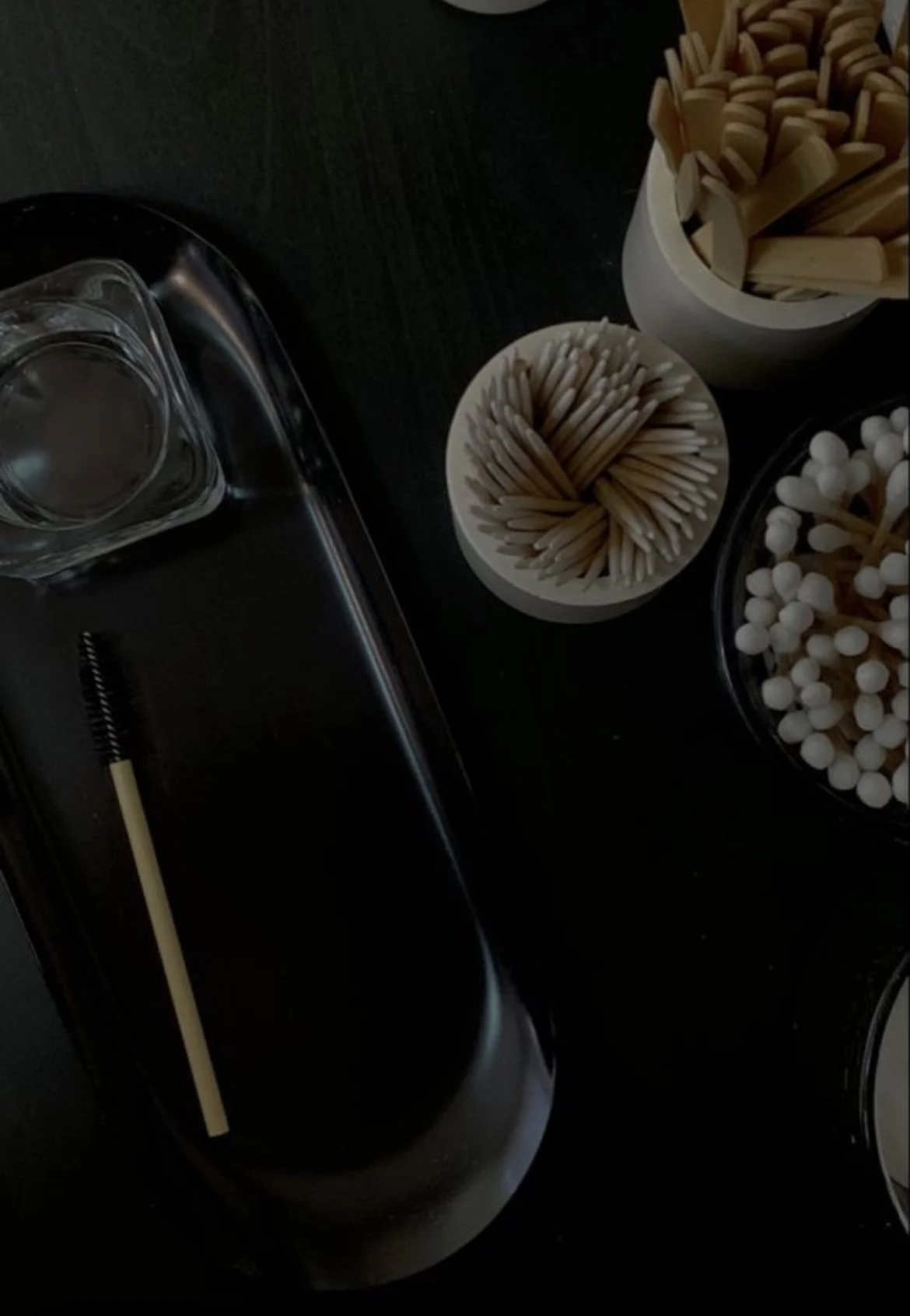 A black tray with a spoolie brush, containers of toothpicks, wooden sticks, and cotton swabs on a dark surface.