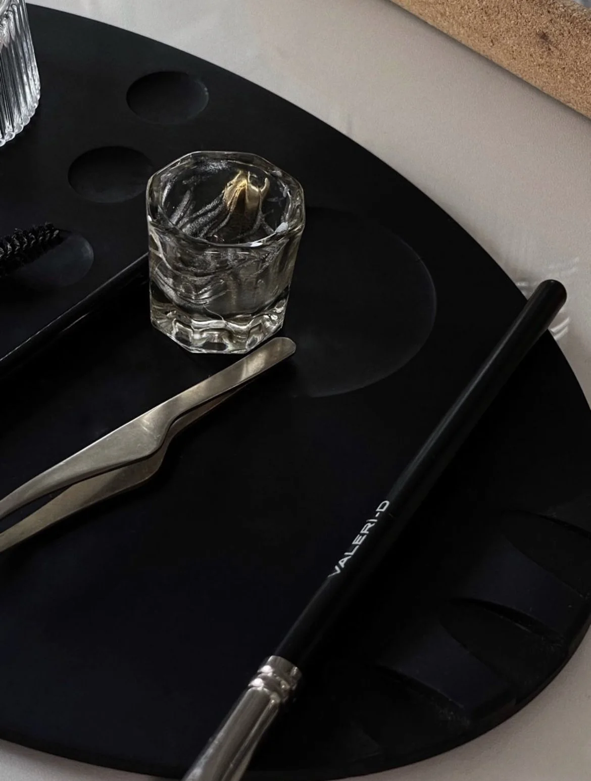 Close-up of a black artist's palette with a glass of water containing a brush, metal tweezers, and a paintbrush with "VALERI-D" imprinted on it.