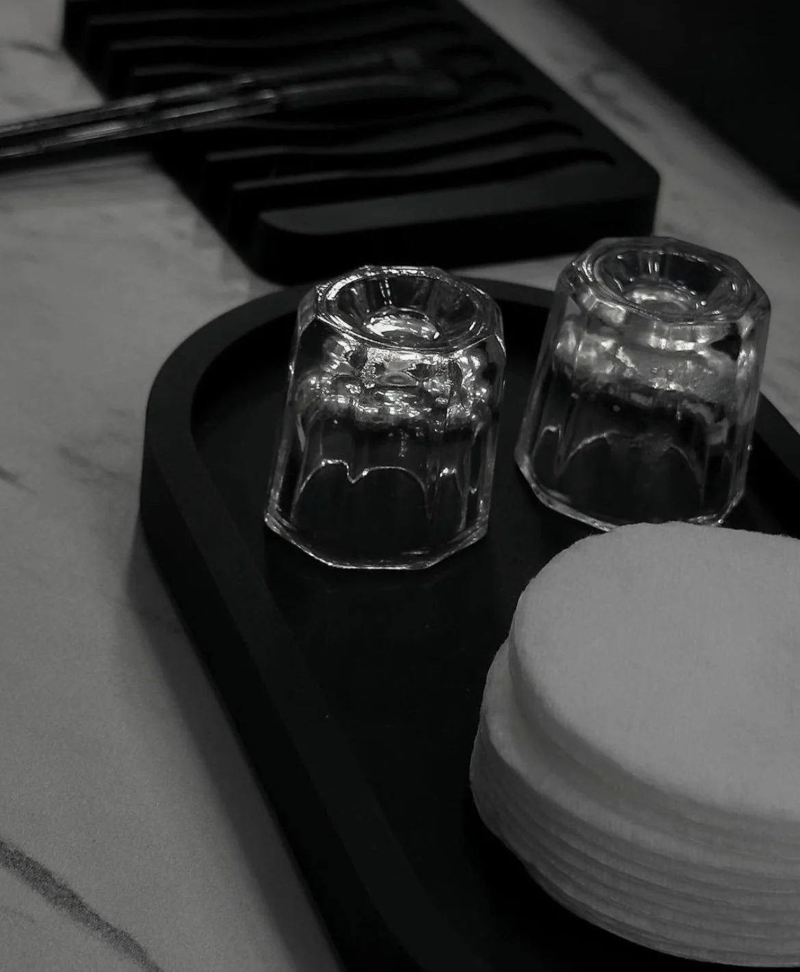 Two upside-down glass cups on a dark tray next to a stack of round cotton pads on a marble surface.