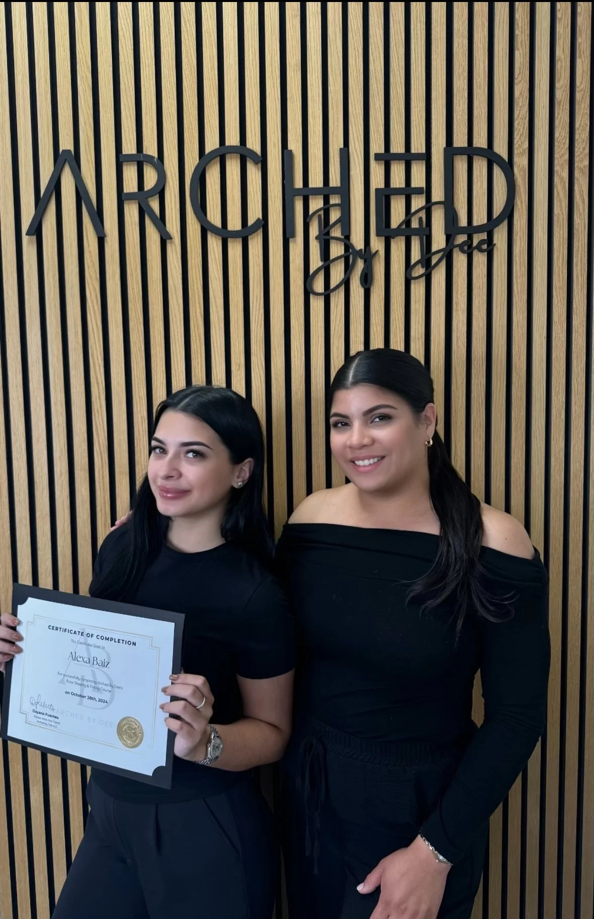 Two women standing in front of a wooden panel wall with the logo "Arched by Dee". One woman holds a certificate of completion. Both are wearing black outfits.
