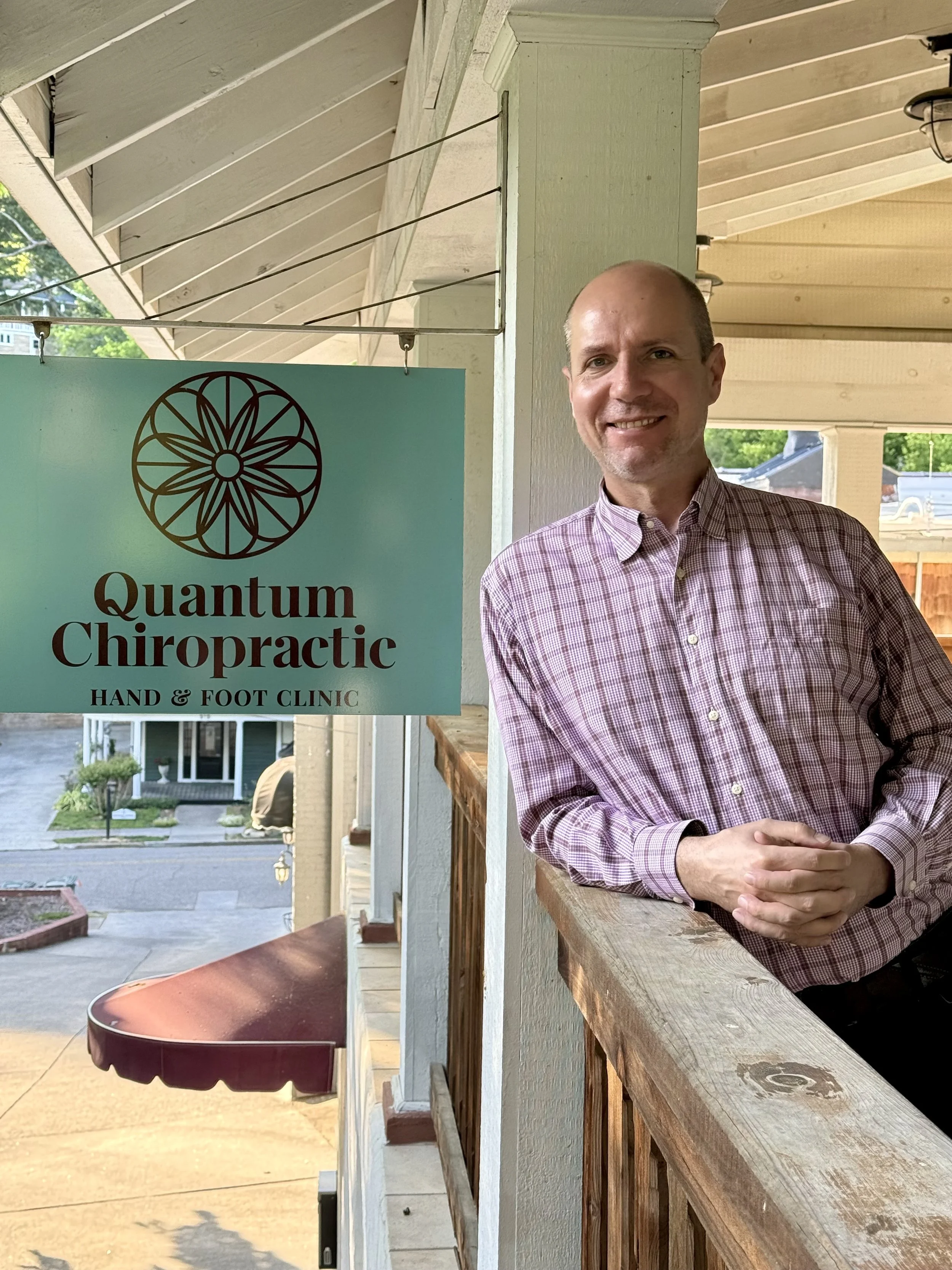 Dr. Bartosz, smiling, wearing a pink checkered shirt, standing outdoors on a porch next to a sign for Quantum Chiropractic Hand & Foot Clinic, with a view of a quiet street and houses in the background.