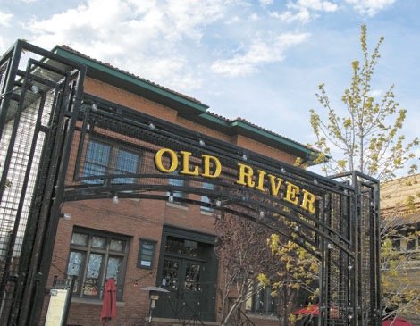 Entrance sign reading 'Old River' on a black metal gate in front of a brick building with windows and a tree with yellow leaves.