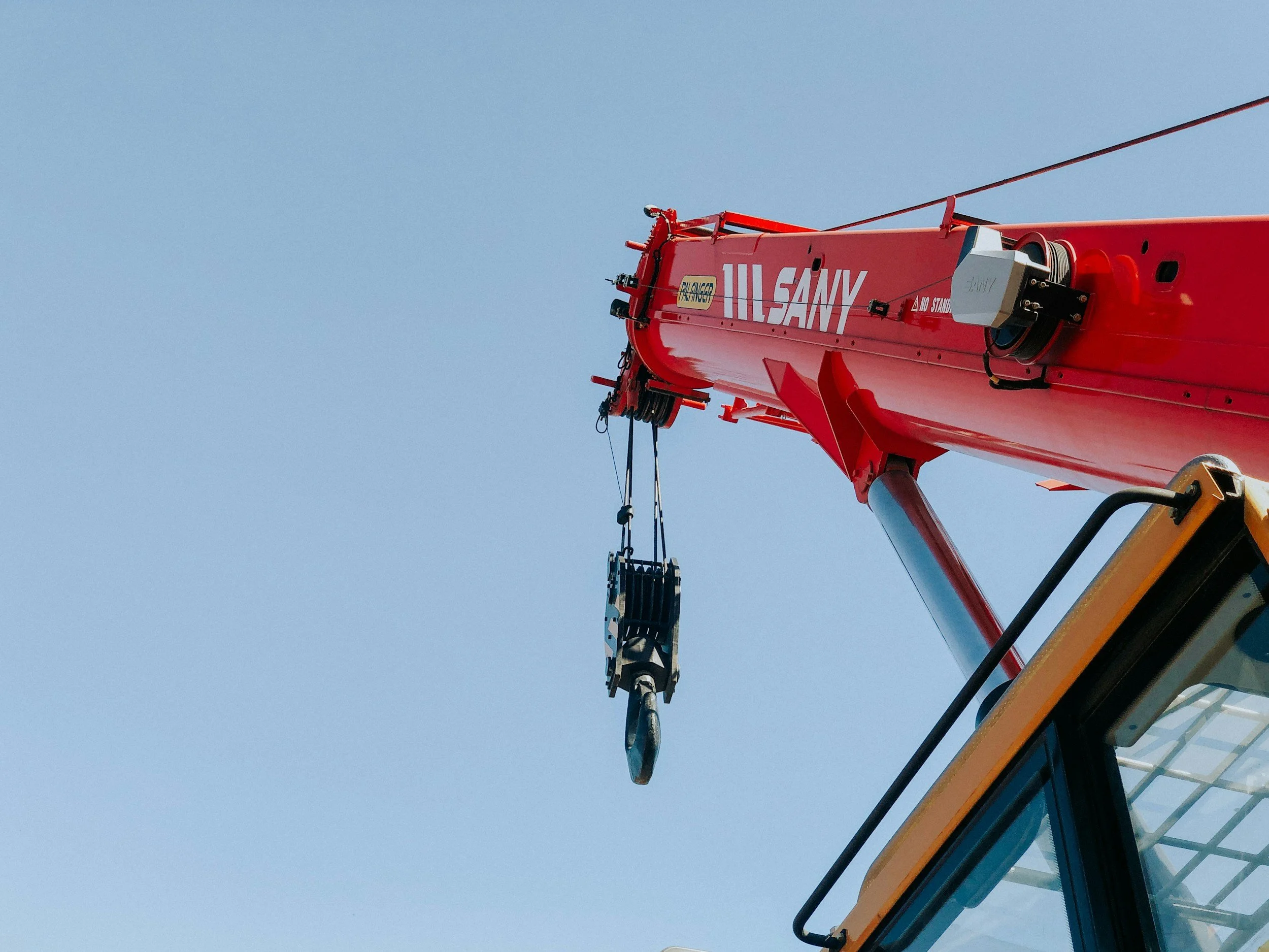 Close-up of a red crane arm with the marking "SANY" and a hook hanging from cables against a clear blue sky.