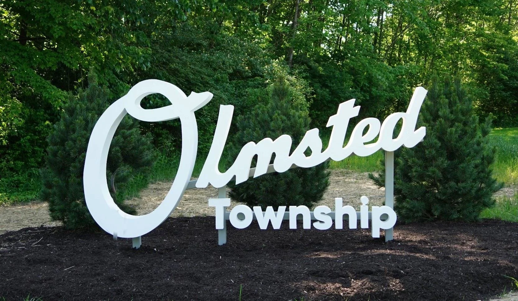 A decorative sign spelling 'Olimsted Township' in white, situated outdoors on a patch of dark soil, with green trees and bushes in the background.