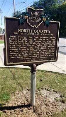 Historic Ohio marker for North Olmsted's first settlement and schoolhouse, with a black background, gold lettering, and green decorative elements at the top corners.